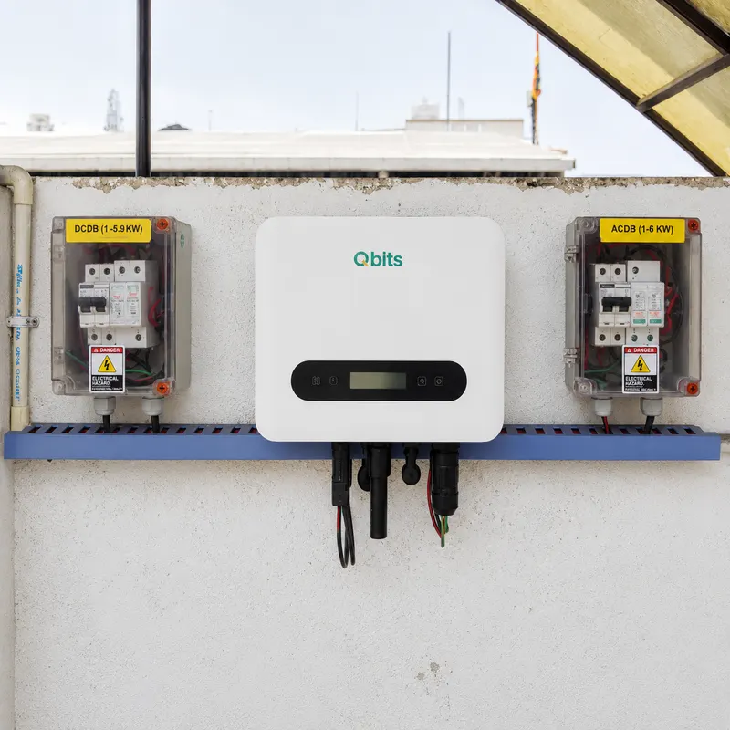 Qbits solar inverter mounted on a white terrace wall under an open pergola, flanked by DCDB and ACDB boxes on a blue cable trunk